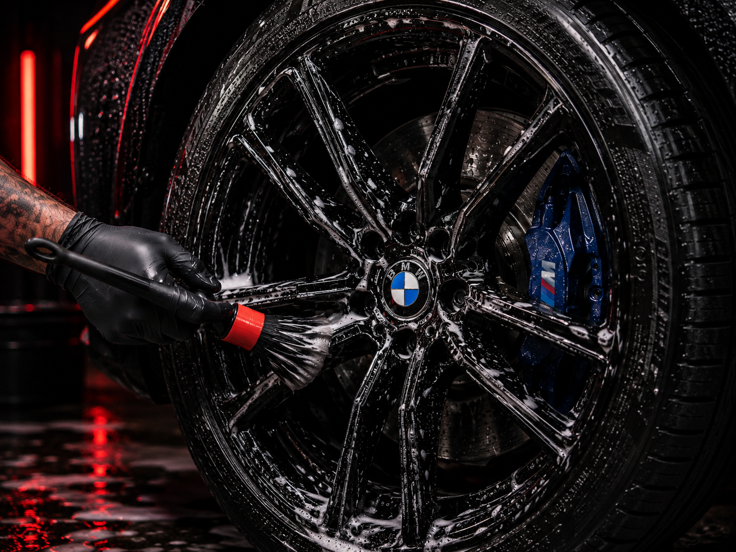 Gloved hand working a detailing brush across a soap-covered black BMW wheel with a blue M brake caliper during the KingWash wheel stage